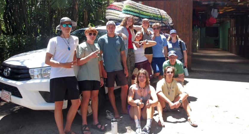 A group of people pose for a photo near a vehicle with surf boards strapped to the top rack. 